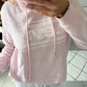Ljusrosa hoodie från Levi's - Säljer en ljusrosa hoodie från Levi's med stort vitt logotryck på bröstet och snörning i huvan. Tröjan har långa ärmar, ribbade muddar och en liten Levi's-tag på ärmen. Perfekt för chill dagar och snygg att matcha med jeans eller mjukis.