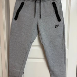Grå Nike Tech Byxor  - Snygga grå Nike Tech mjukisbyxor från Nike med svarta dragkedjor på fickorna och justerbar snörning i midjan. Byxorna har en slim passform och är tillverkade i mjukt bomullsmaterial. Perfekta för chill eller träning, med klassisk Nike-logga på låret.
