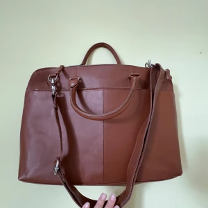 Brun datorväska från Sandqvist - Snygg brun datorväska från Sandqvist i skinn med stilren design. Väskan har både handtag och justerbar axelrem, samt flera fack inuti för laptop och tillbehör. Perfekt för dig som vill ha en klassisk och praktisk väska med dragkedja.