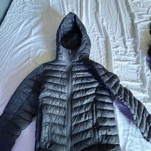 Blå dunjacka Peak Performance 130 - Säljer en blå dunjacka från Peak Performance med huva och dragkedja framtill. Jackan har ett quiltat mönster och är perfekt för kyliga dagar. Snygg, sportig look och bekväm passform. Logga på bröstet och två sidofickor med dragkedja.