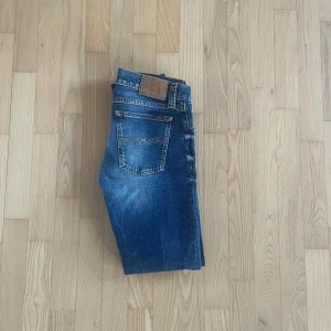 Blå jeans från Nudie Jeans - Snygga blå jeans från Nudie Jeans med klassisk femficksdesign och coola slitningar. Jeansen har en rak passform och är tillverkade i mjukt denimtyg med kontrastsömmar och läderpatch bak i midjan. Perfekta för dig som gillar en avslappnad och stilren look.
