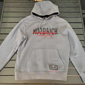 Grå hoodie från Hoodrich med tryck - Säljer en grå hoodie från Hoodrich i storlek L. Tröjan har svart och rött tryck på bröstet, snörning i huvan och en stor magficka. Insidan av huvan är svart och det finns en patch längst ner på fickan. Perfekt för chill eller streetwear.