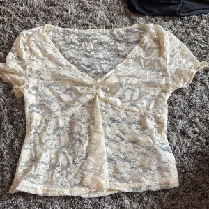 Beige spetsblus från SHEIN - Säljer en beige, kortärmad spetsblus från SHEIN i storlek S. Blusen har v-ringning och är gjord i ett transparent spetsmaterial med blommigt mönster. Den har en snygg twist-detalj framtill som ger extra stilpoäng. Perfekt för dig som gillar romantiska vibes.