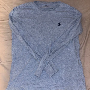 Blå långärmad tröja från Polo Ralph Lauren - Säljer en stilren blå långärmad tröja från Polo Ralph Lauren med klassisk rund hals och mörkblå broderad logga på bröstet. Tröjan är gjord i mjuk bomull och har en normal passform som är skön att bära. Perfekt för dig som gillar enkel och snygg design.