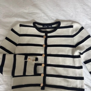 Randig kofta - Snygg kofta från French Connection i storlek S. Den är vit med svarta horisontella ränder och har guldfärgade knappar framtill. Koftan är stickad och har långärmade ärmar, perfekt för att ge din outfit en fräsch och klassisk vibe.