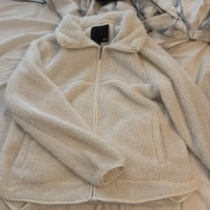 Vit teddyjacka från lager 157 XS - Mysig vit teddyjacka från lager 157 i storlek XS. Jackan har fluffigt fleece-material, hel dragkedja framtill och två sidofickor. Perfekt för kalla dagar när du vill vara både bekväm och snygg. Hög krage för extra värme och stil.