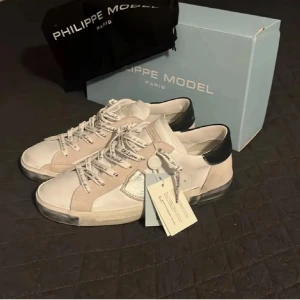 Säljer mina helt nya Philip modells nu ny pris 3300kr - Snygga vita sneakers från Philippe Model med beige och silverdetaljer samt svart häl. Skorna har snörning med logotyp på skosnörena och en rund tå. Materialmix av skinn och mocka ger en lyxig känsla. Perfekt för dig som gillar stilrena och trendiga sneakers.
