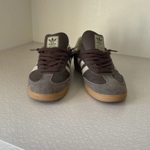 Adidas Samba sneakers brun/vit - Adidas Samba sneakers i mörkbrunt och vitt med klassiska tre ränder på sidan. Ovandel i skinn och mocka, gummisula i beige. Låg modell med rund tå och snörning. Perfekt för dig som gillar retrostil och sportig vibe. Box finns