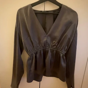 Svart satinblus med v-ringning - Snygg grå blus från Gina Tricot i glansigt satinmaterial. Blusen har en djup v-ringning, långärmade ärmar och ett smickrande rynkat parti under bysten som ger en cool siluett. Perfekt för att lyfta din outfit med lite extra stil.