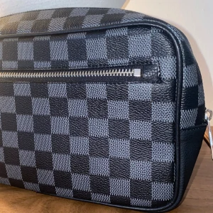 Rutig svart axelväska i skinn - Snygg axelväska i svart och grått med klassiskt rutigt mönster. Väskan har en praktisk dragkedja framtill och är tillverkad i tåligt skinn. Perfekt för dig som vill ha en stilren och ikonisk accessoar.