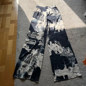 Helt oanvända wide satinbyxor från H&M   - Säljer ett par svarta och vita wide byxor från H&M i storlek 34. De har ett coolt grafiskt mönster med stadsmotiv och en elastisk midja för extra komfort. Materialet är mjukt och lätt, perfekt för dig som gillar statement-plagg med avslappnad vibe.