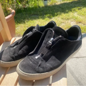 Svarta mockasneakers med dragkedja från Axel Arigato - Svarta sneakers ifrån Axel Arigato i mjuk mocka med cool dragkedja framtill istället för klassisk snörning. Snygg kontrast med vit platt sula och detaljer i svart skinn på hälen. Perfekt för dig som gillar minimalistisk stil med en twist. Säljer för dom inte passade mig