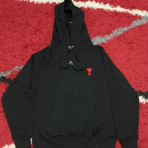 Svart hoodie från AMI PARIS med rött hjärta - Svart hoodie från AMI med klassisk känguruficka och snörning i huvan. På bröstet finns ett broderat rött hjärta med bokstaven A. Tröjan har långa ärmar och ribbade muddar. Perfekt för chill dagar och streetwear-stil.