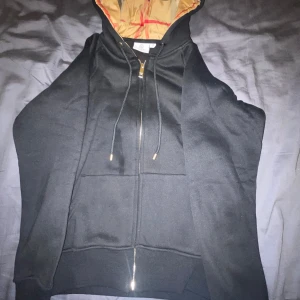 Svart Burberry hoodie med dragkedja - Säljer en svart hoodie från Burberry med klassisk rutmönstrad huva i beige och rött. Tröjan har hel dragkedja, snörning vid huvan och två fickor framtill. Perfekt för dig som vill ha en stilren och lyxig look med ikonisk Burberry-touch.