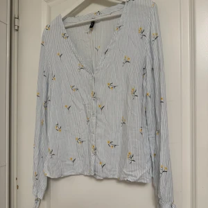 Blåvit blommig skjorta från Divided - Snygg skjorta med ljusblå och vita ränder, dekorerad med små gula blommor. V-ringad med knappar framtill och långa ärmar med knyt vid ärmslut. Luftigt material i bomull, perfekt för vår och sommar. Fräsch och trendig look! Osb storlek  L men väldigt liten i storleken.