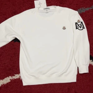 Vit sweatshirt från Moncler med logga - Snygg vit sweatshirt från Moncler med rund hals och långa ärmar. Tröjan har Moncler-logga på bröstet och ett stort svart M-tryck med logga på ena ärmen. Mjuk och skön bomullskvalitet, perfekt för chill eller streetwear.