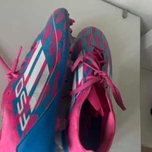 Adidas F50 blå och rosa fotbollsskor - Säljer ett par Adidas F50 fotbollsskor i blått och rosa med vita ränder. Skorna har snörning och en mönstrad ovandel med grafiska detaljer. Sulan är blå med rosa dobbar, perfekt för gräsplan. Lätta och snygga skor som sticker ut på planen.