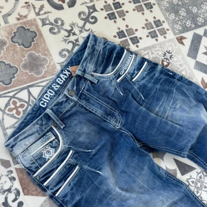 Blå jeans från Cipo & Baxx med vita detaljer - Snygga blå jeans från Cipo & Baxx med coola vita detaljer och kontrastsömmar. Modellen har flera fickor med dragkedjor och logotyp på både linning och bakficka. Jeansen har en modern passform och slitningar för en streetig vibe. Storlek 30/32