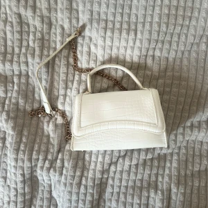 Vit handväska med kedja - Snygg vit handväska med krokodilpräglad yta och guldfärgad kedja som axelrem. Väskan har ett stilrent och kompakt format med ett handtag upptill. Perfekt för att lyfta din outfit med en clean och trendig touch.