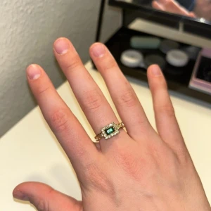 Guldring med grön sten och kristaller - Snygg guldring med en rektangulär grön sten i mitten, omgiven av små glittrande kristaller. Ringen har en elegant och lyxig känsla, perfekt för dig som gillar att sticka ut med dina accessoarer. Passar både till jeans och klänning.