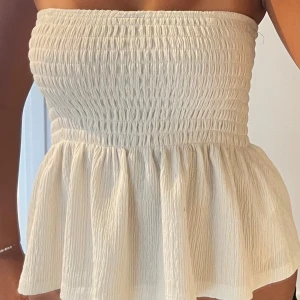 Vit bandeau tubtopp med volang - Supersöt bandeau tubtopp i ljusvit färg med smockad överdel och volang nedtill. Toppen är ärmlös och har en luftig, flowy passform som ger en chill vibe. Perfekt för varma dagar och sommarhäng.