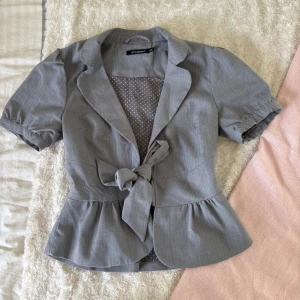 Grå topp med knyt - Snygg grå topp/kavaj från Atmosphere i storlek 36. Kortärmad modell med volang nedtill och ett brett knytband i midjan. Insidan har ett prickigt foder.💞