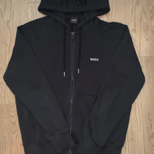 Hugo boss zip hoodie  - Svart hoodie från BOSS med dragkedja, huva och snörning. Framsidan har en liten vit BOSS-logga och baksidan har coola grafiska tryck i gråskala med månlandskapsmotiv. Mjuk bomullskänsla och praktiska fickor framtill. Perfekt för chill dagar.