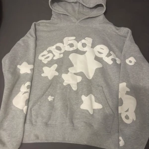 Sp5der hoodie  - Grå hoodie från Sp5der i storlek M, tillverkad i 100% bomull. Tröjan har en stor huva och coola vita stjärnor och texttryck över hela framsidan och ärmarna. Perfekt för dig som gillar streetwear och vill sticka ut med ett unikt mönster.