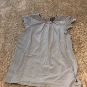 Ljusgrå blus med spets från Jeannie - Supersöt ljusgrå blus från Jeannie med korta ärmar i spets och rund halsringning med knapp i nacken. Spetsdetaljerna har ett snyggt virkat mönster som ger en romantisk vibe. Perfekt att matcha med jeans eller kjol för en chill look.