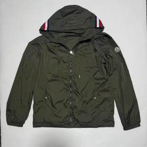 Moncler grön vindjacka med huva - Snygg grön vindjacka från Moncler med huva och coola röd-blå-vita detaljer. Jackan har dragkedja framtill, två fickor med knappar och elastiska muddar. Materialet är lätt och syntetiskt, perfekt för blåsiga dagar. Moncler-logga på ärmen ger extra stilpoäng.