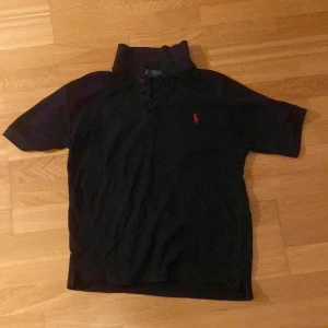 Svart Polo Ralph Lauren t-shirt M - Svart t-shirt från Polo Ralph Lauren i storlek M. Klassisk pikémodell med korta ärmar, krage och knappar framtill. Röd broderad logga på bröstet. Tillverkad i mjuk bomull som är skön mot huden. Perfekt för dig som gillar stilrena och tidlösa plagg.