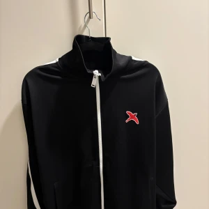 Svart zip hoodie med vita ränder - Svart hoodie med vit dragkedja och vita ränder längs ärmarna. På bröstet sitter en röd broderad fågel. Hög krage och långa ärmar ger en sportig vibe. Perfekt för chill eller träning. Materialet känns som polyester och är mjukt mot huden.