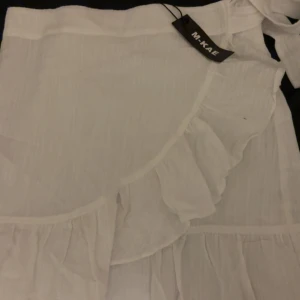 Vit omlottkjol med volanger från M-KAE - Supersöt vit omlottkjol från M-KAE med volangdetaljer och lätt, luftig känsla. Kjolen har knytband i midjan och är perfekt för varma dagar. Den är kort och har en feminin, flowy stil som ger en fräsch vibe. Den är stum i midjan, därför ja säljer då den e för tajt på mig så kan inte dra upp dragkedjan. Kan ta ljusa bilder om de önskas💕