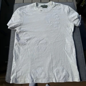 Vit Denim Tears t-shirt med blommor - Säljer en vit t-shirt från Denim Tears med subtila blommiga broderier på framsidan. T-shirten har rund hals, korta ärmar och är gjord i mjuk bomull. Perfekt för dig som gillar clean stil med unika detaljer.