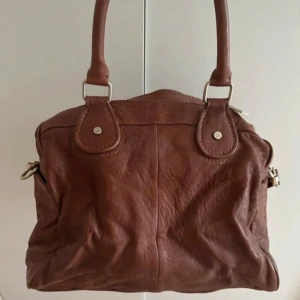 Brun väska från Paul & Joe - Snygg brun väska  från Paul & Joe i mjukt skinn med silverfärgade detaljer och dragkedja. Väskan har både handtag och justerbar axelrem, samt praktiska fack och logga på framsidan. Perfekt storlek för allt du behöver och stilren design