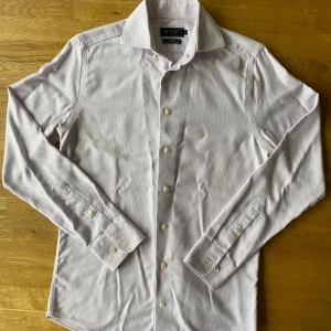 Ljusrosa slim fit skjorta från Riley - Snygg ljusrosa skjorta från Riley i slim fit-modell med diskret mönster och klassisk krage. Skjortan har långa ärmar, knappar framtill och är perfekt för dig som gillar stilrena och moderna plagg. Materialet känns mjukt och lätt mot huden.