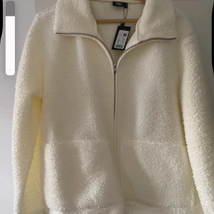 Vit teddyjacka med dragkedja - Mysig vit teddyjacka från 157 med hög krage och hel dragkedja framtill. Jackan har två stora fickor och är tillverkad i fluffigt fleece-material som ger en cool och avslappnad vibe. Perfekt för att hålla värmen under kyliga dagar.