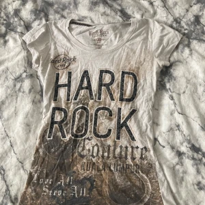 Hard Rock Couture t-shirt XS - Cool t-shirt från Hard Rock Couture i storlek XS. T-shirten är vit med stora tryck i svart och brunt framtill, och har korta ärmar samt rund hals. Materialet är mjuk bomull blandat med polyester, vilket gör den skön och lätt att bära. Perfekt för dig som gillar statement-plagg.