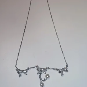 Sött halsband med rosetter och pärlor - Ett silvrigt halsband med tre glittriga rosetter som hängsmycken, dekorerade med små stenar. Två vita pärlor hänger från mittenrosetten och ger extra detalj. Kedjan är tunn och har ett klassiskt lås. Perfekt för dig som gillar gulliga och eleganta smycken.