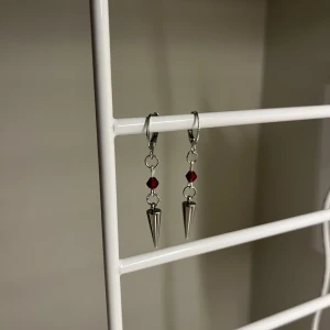 Silvriga örhängen med röda pärlor -  I rostfritt stål och handgjort. Coola silvriga örhängen med hängande konformade detaljer och röda pärlor. Perfekt för dig som gillar edgy och unik stil. Snyggt att matcha med både streetwear och mer dressade outfits.