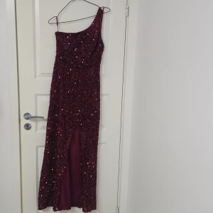 Vinröd glittrig långklänning med slits - Säljer en vinröd långklänning med glittriga paljetter över hela klänningen. Den har en snygg oneshoulder-design med en smal axelrem och en hög slits framtill. Perfekt för dig som vill sticka ut och glänsa på bal eller fest!