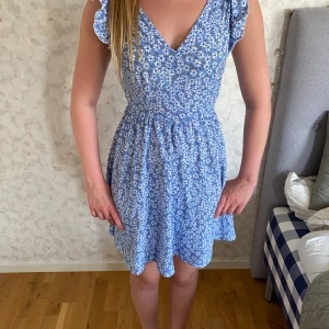 Blommig blå kortklänning från SHEIN - Supersöt blå klänning med vita blommor från SHEIN. Kort modell med volangdetaljer på axlarna, v-ringad framtill och öppen rygg med knytband. Perfekt för sommarens alla tillfällen! Lätt och skön i bomullsmaterial.