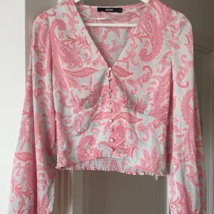 Rosa från Bik Bok - Supersöt blus från Bik Bok i storlek S med paisleyinspirerat mönster i rosa. Blusen har croppad passform, långa ärmar och v-ringad hals. Framtill finns klädda knappar och smockad nederkant som ger en snygg siluett. 