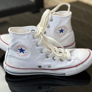 Vita Converse Chuck Taylor All Star - Klassiska vita Converse Chuck Taylor All Star high tops med röd och blå rand runt sulan. Jättefint skick. Stl 35. 