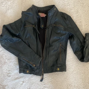 Svart skinnjacka från Saint Tropez - Snygg svart skinnjacka från Saint Tropez med coola detaljer som dragkedjor på fickor och ärmar, samt knappar vid nederkant. Jackan har en klassisk bikerstil med ståkrage och figurnära passform. Perfekt för dig som vill ha en edgy look.