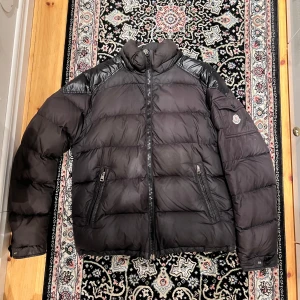 Svart Moncler dunjacka herr Chevalier storlek 4/L - Svart dunjacka från Moncler med glansiga detaljer på axlarna och insidan. Jackan har hög krage, två fickor med dragkedja och en ficka med Moncler-logga på ärmen. Perfekt för kalla dagar och har en riktigt snygg streetstil.