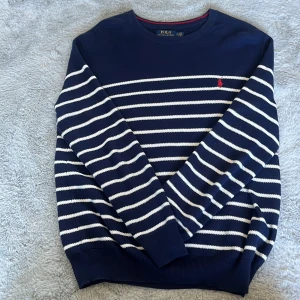 Marinblå randig tröja från Polo Ralph Lauren - Säljer en marinblå tröja från Polo Ralph Lauren med vita horisontella ränder och röd broderad logga på bröstet. Tröjan har rund halsringning och långa ärmar. Perfekt för en stilren och klassisk look.