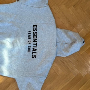 Grå Essentials Fear of God hoodie - Säljer en grå hoodie från Essentials Fear of God. Perfekt för en avslappnad och trendig stil. Tröjan är helt oanvänd, den är alldeles för stor för mig för att använda. 