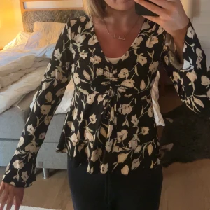 Svart blommig blus med v-ringning - Säljer en svart blus med vita blommor och v-ringning. Blusen har långa ärmar och lätt utsvängd nederkant. Snygg passform med knytning framtill som ger en feminin siluett.