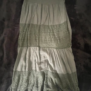 Vit långkjol med spetsdetaljer - Säljer en romantisk vit långkjol med breda spetsinslag och volanger. Kjolen har en luftig och flowy känsla, perfekt för varma dagar. Passar dig som gillar boho-stil och vill ha något extra till sommargarderoben. Har aldrig använt den och köpte den för 280kr